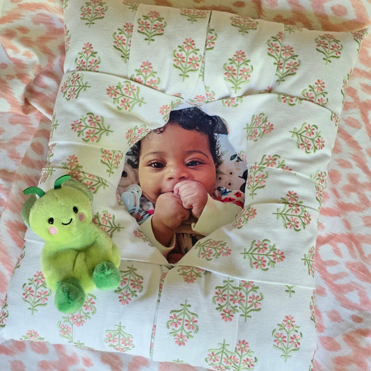 An overstuffed picture frame with an embroidered pink and green floral pattern on a white background, featuring a custom photo cutout in the center showing a smiling baby.There's a green baby's stuffie attached at the right bottom of the frame. The frame is displayed on a light pink floral background.