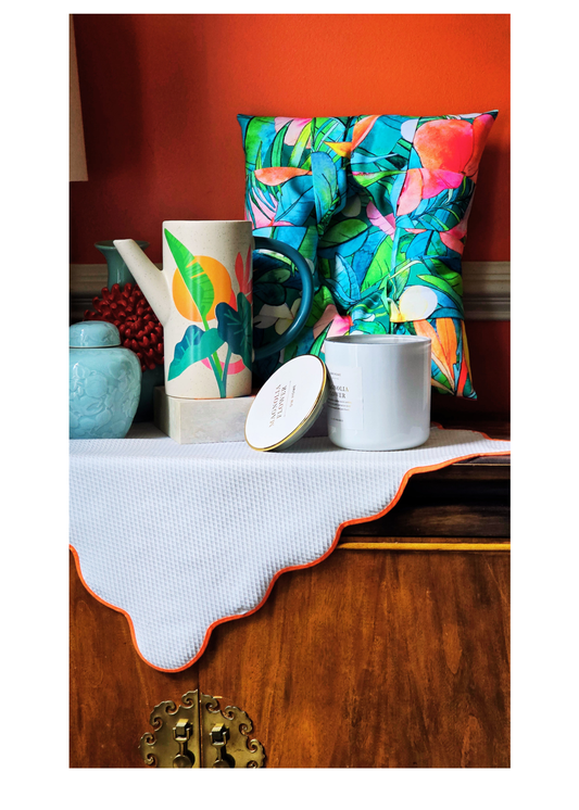 A styled tabletop arrangement featuring a colorful tropical leaf print overstuffed picture frame, a matching patterned ceramic pitcher, a white candle with a gold lid, and a white textured cloth with an orange scalloped edge, set against a warm orange wall and wooden cabinet.