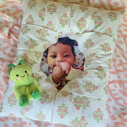 An overstuffed picture frame with  an embroidered pink and green floral pattern on a white background, featuring a custom photo cutout in the center showing a smiling baby.There's a green baby's stuffie attached at the right bottom of the frame. The frame is displayed on a light pink floral background.