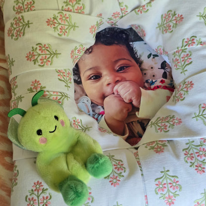 A close-up of a baby's face in a white-based floral overstuffed picture frame, with a green plush toy attached as embellishment.