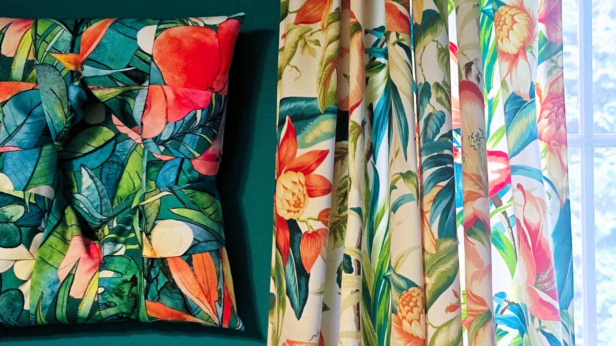 A full front view of an overstuffed picture frame in tropical fabric with bright , yellows, blues and greens; shown next to a window.with tropical floral curtains with a vibrant multi-color print on white featuring green and blue leaves, red and orange flowers.