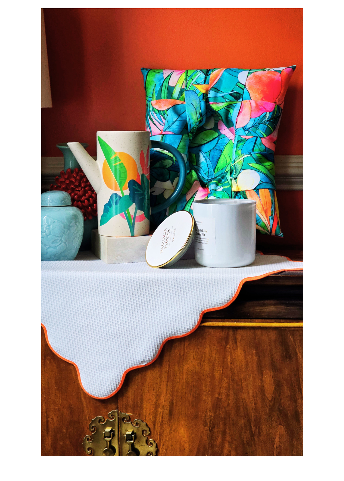 A styled tabletop arrangement featuring a colorful tropical leaf print overstuffed picture frame, a matching patterned ceramic pitcher, a white candle  with a gold lid, and a white textured cloth with an orange scalloped edge, set against a warm orange wall and wooden cabinet.