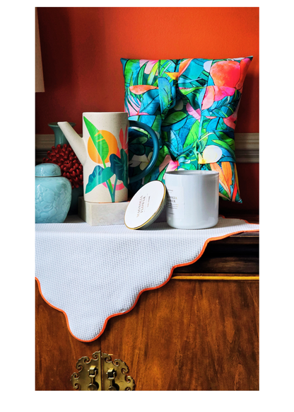 A styled tabletop arrangement featuring a colorful tropical leaf print overstuffed picture frame, a matching patterned ceramic pitcher, a white candle  with a gold lid, and a white textured cloth with an orange scalloped edge, set against a warm orange wall and wooden cabinet.