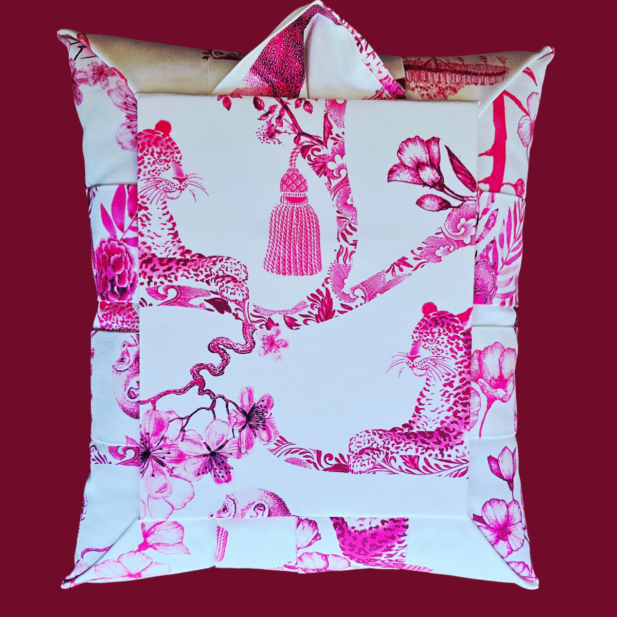 The back view of an overstuffed picture frame with a white base and a vibrant pink and magenta print of leopards, branches, a tassel detail and floral motifs, set against a solid dark red background.